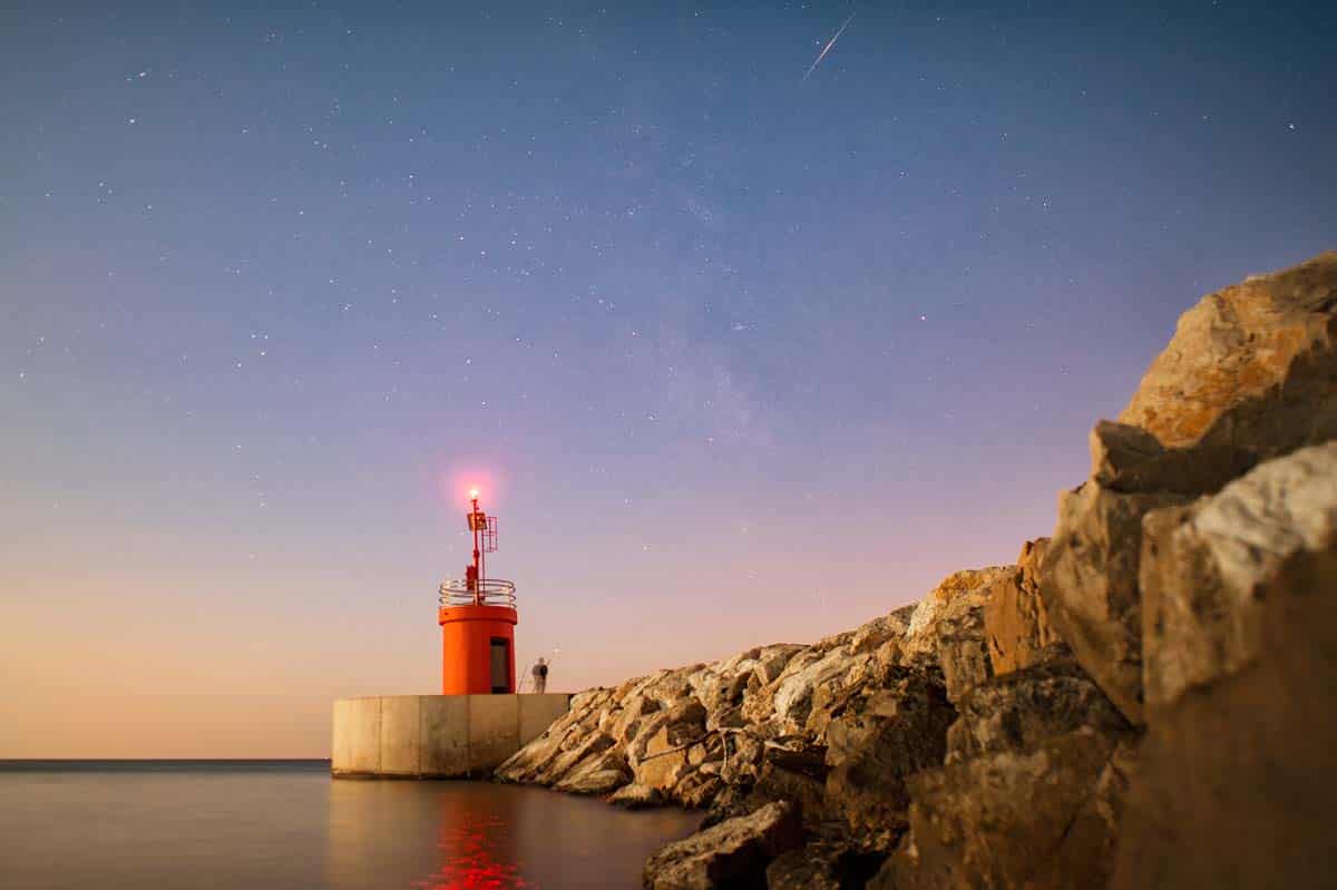 Seascape of a small lighthouse with their reflections on the sea at sunset. A man is fishing under the starry sky. Photo taken at sunset in Sabaudia beach, Italy